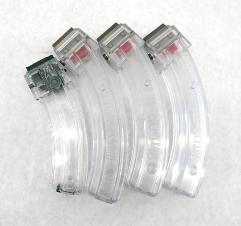 Four clear, curved firearm magazines with visible ammunition, resting on a white surface.