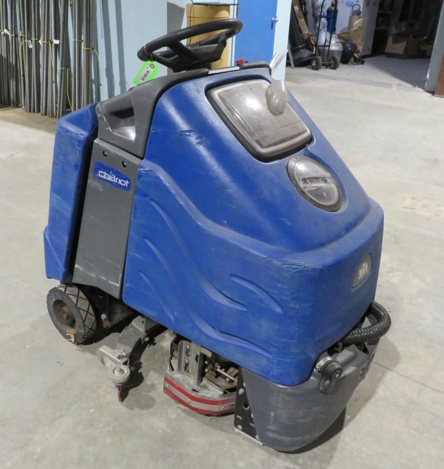 Blue and gray walk-behind floor scrubber machine with a steering wheel in a warehouse.