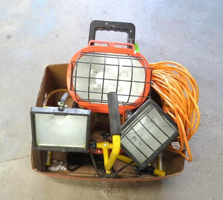 A cardboard box holds orange work lights, power cord, and a yellow stand.