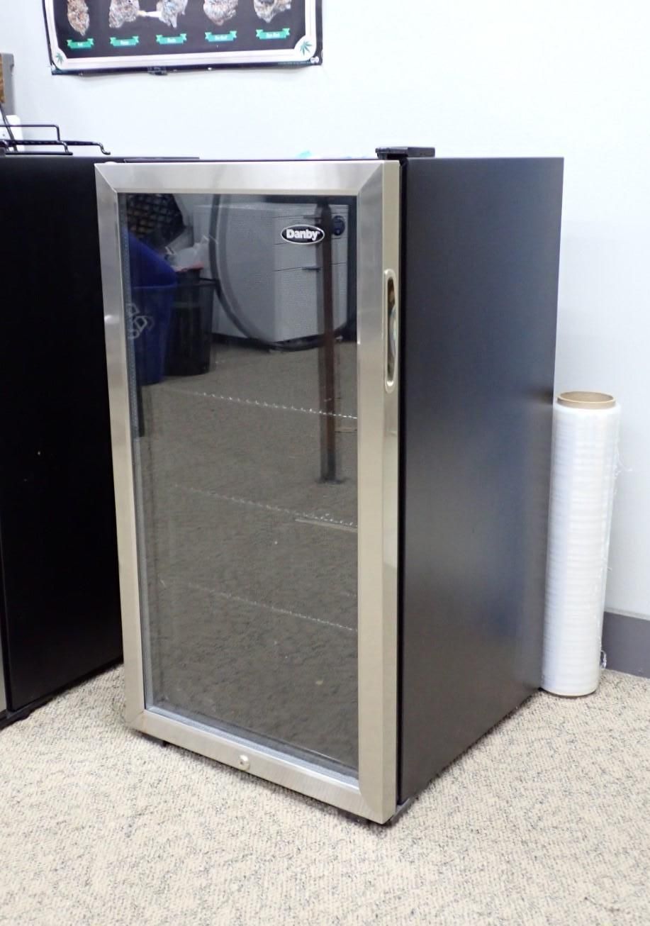 Mini fridge with glass door and silver handle. Black side, white wall, roll of plastic wrap.