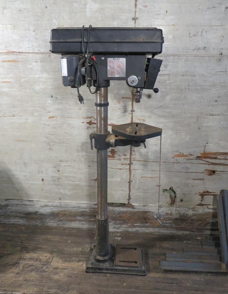 Black drill press machine standing against a light-colored, mottled wall.