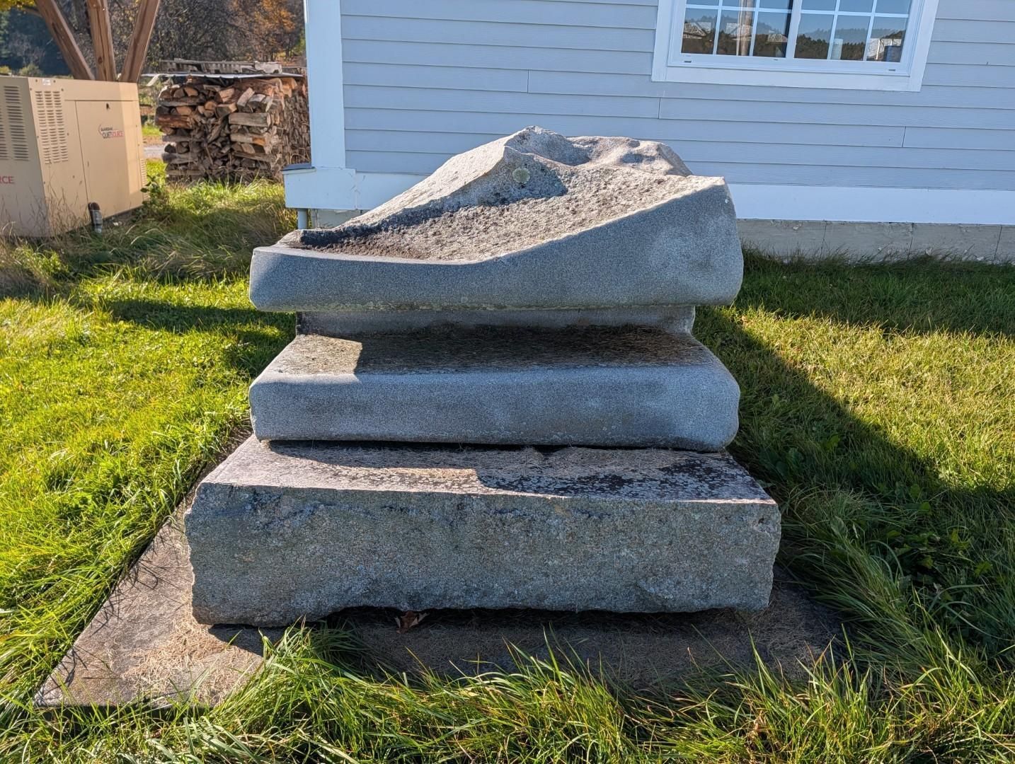 Piled gray stone slabs on grass beside a white house wall