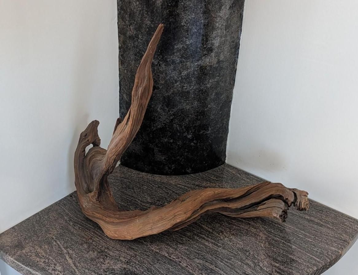 Twisted wooden sculpture leaning against a dark cylindrical post on a gray carpeted corner.
