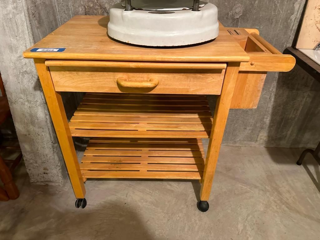 Wooden kitchen cart with a drawer, two slatted shelves, and a cutting board on top. It sits on wheels.