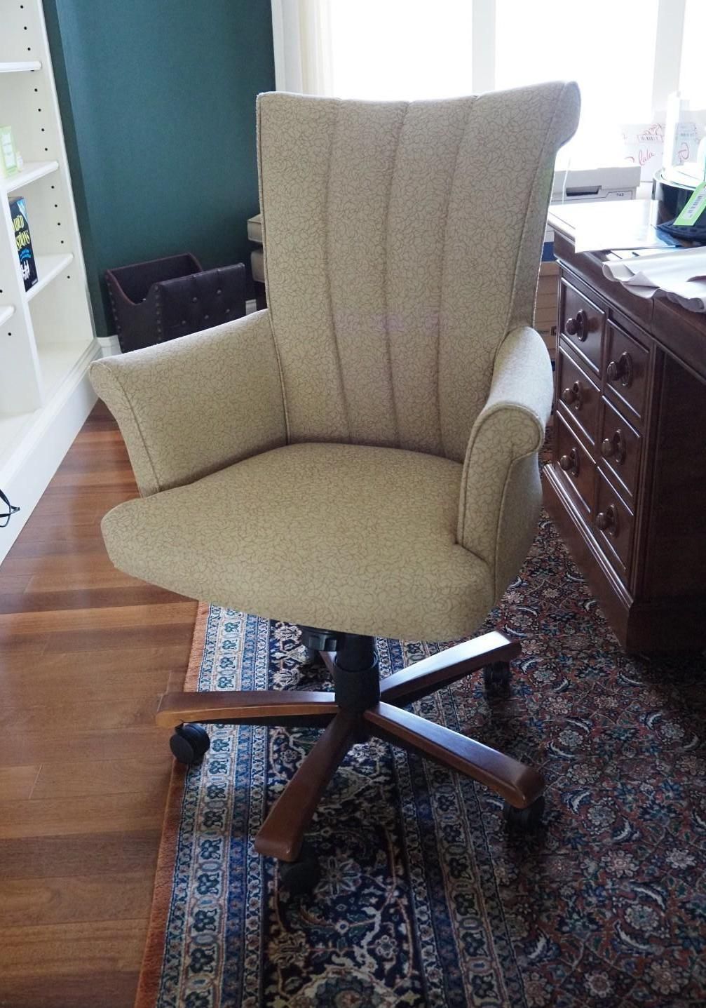 Cream-colored upholstered office chair with wooden base, set on patterned rug, near desk and bookcase.