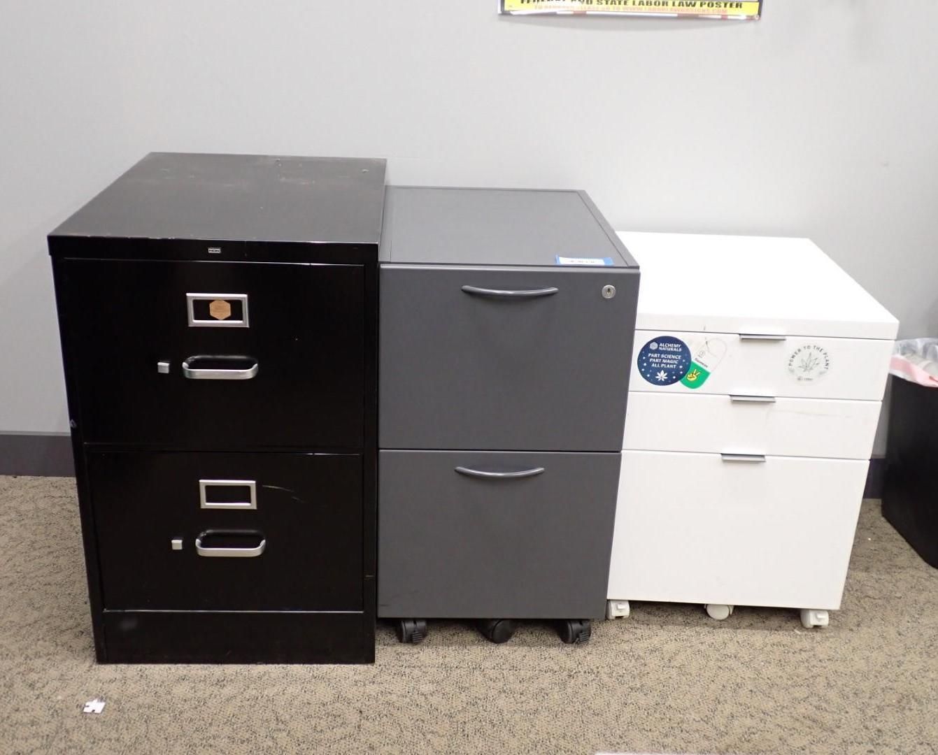 Three file cabinets, black, gray, and white, sit side-by-side on a carpeted floor.