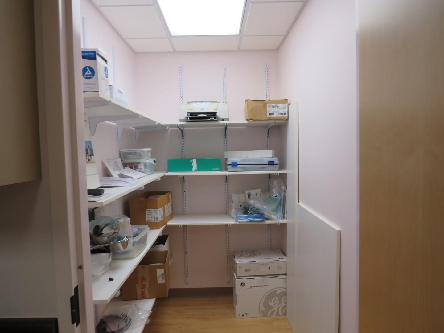 Small storage room with white shelves holding boxes and supplies; pink walls, fluorescent lighting.