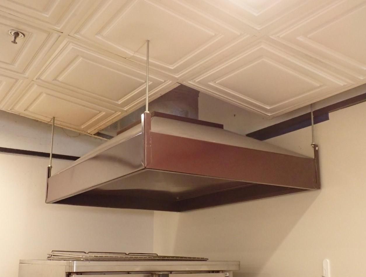 Stainless steel range hood suspended from a white, patterned ceiling, above kitchen equipment.