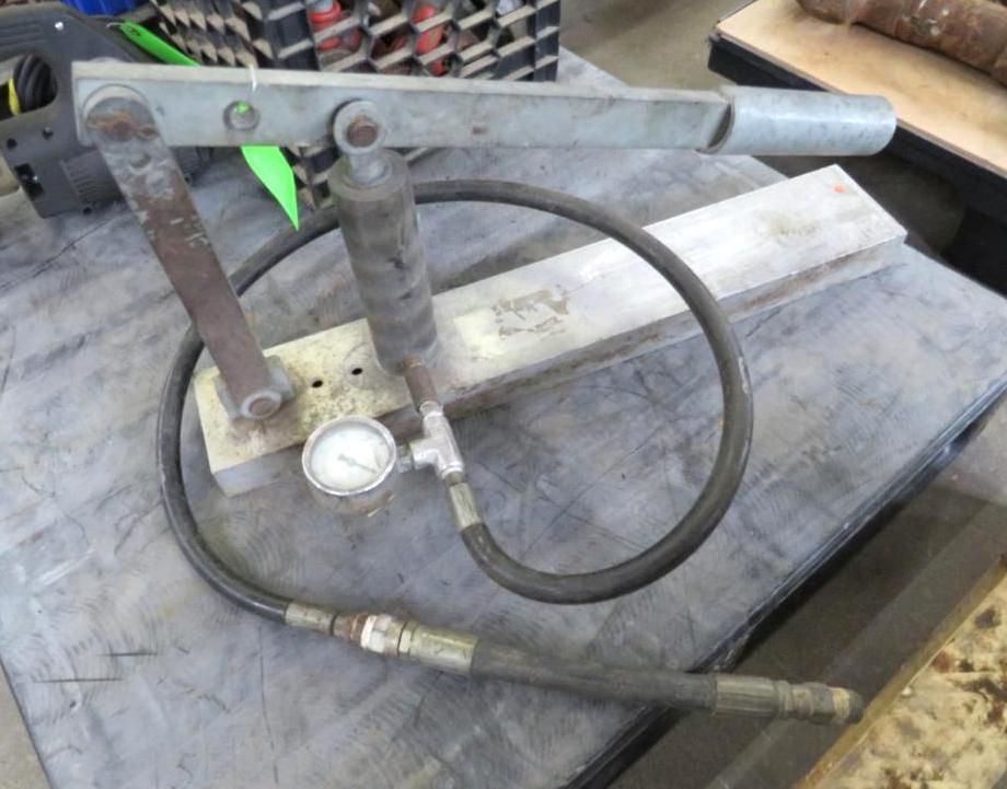 Hand-operated hydraulic pump with gauge and hose on a table.