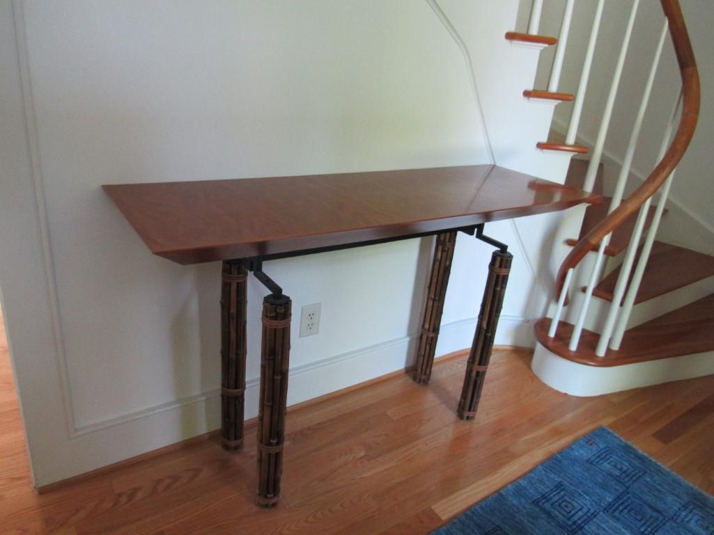 A long, brown table with decorative legs against a white wall and a spiral staircase.