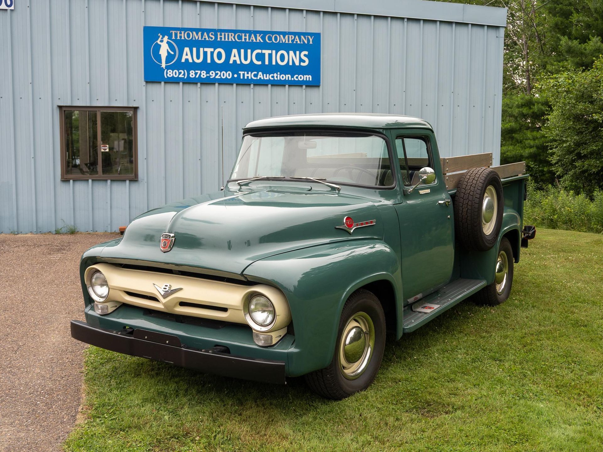 Green vintage Ford pickup truck parked in front of a blue sign for an auto auction.