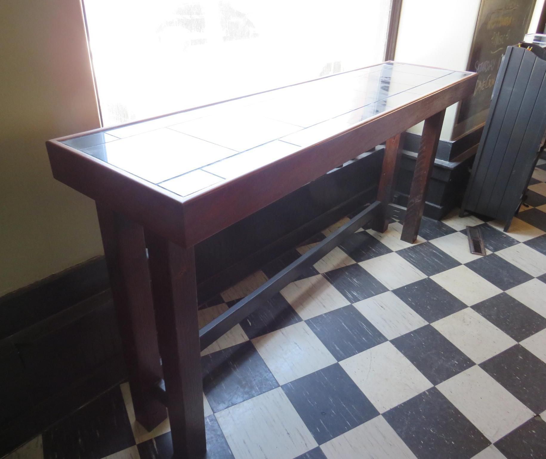 Long, dark wood console table with tiled top, in front of a window, on a checkered floor.