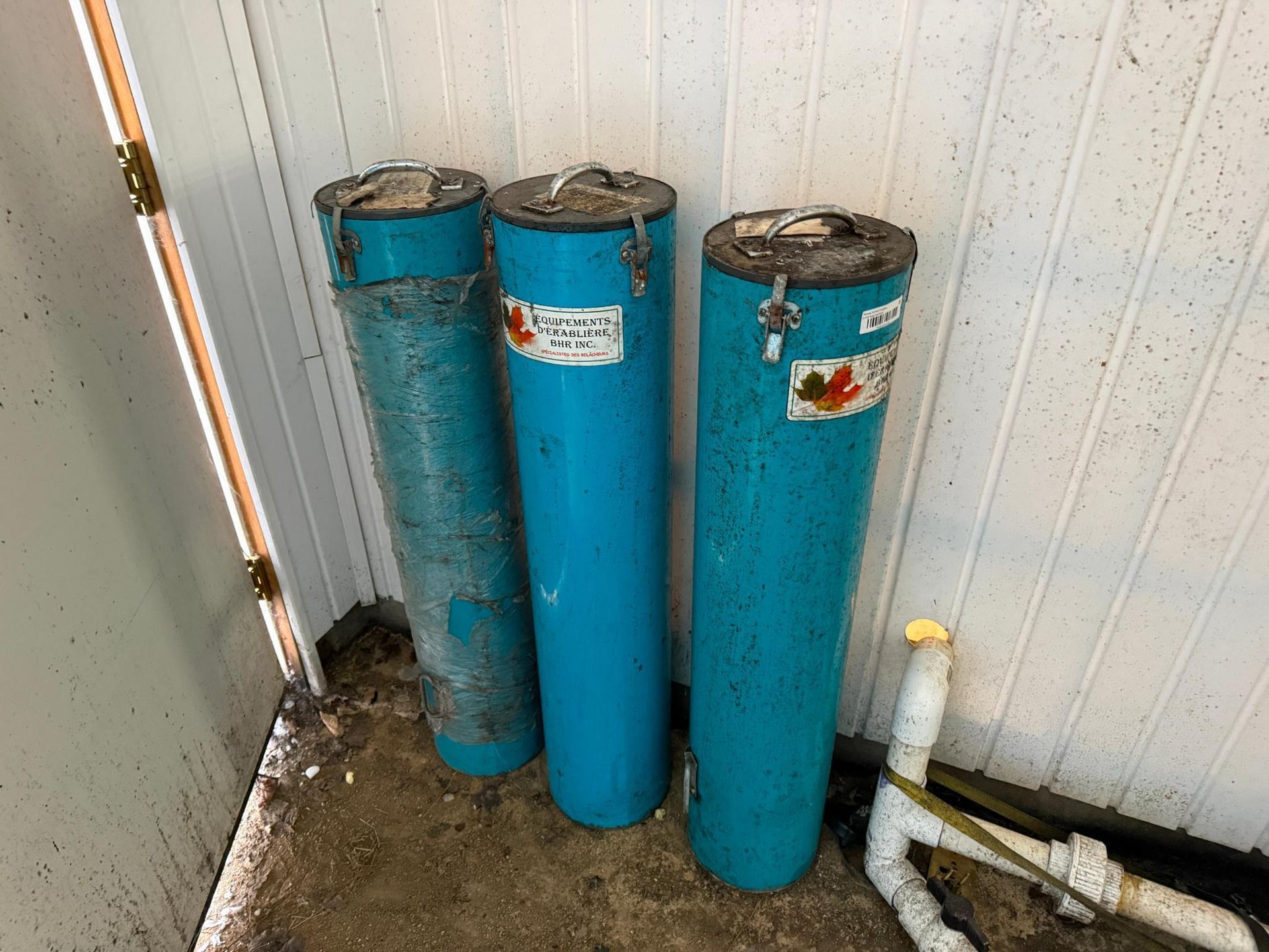 Three tall, blue cylindrical tanks stand against a white wall and a white pipe.