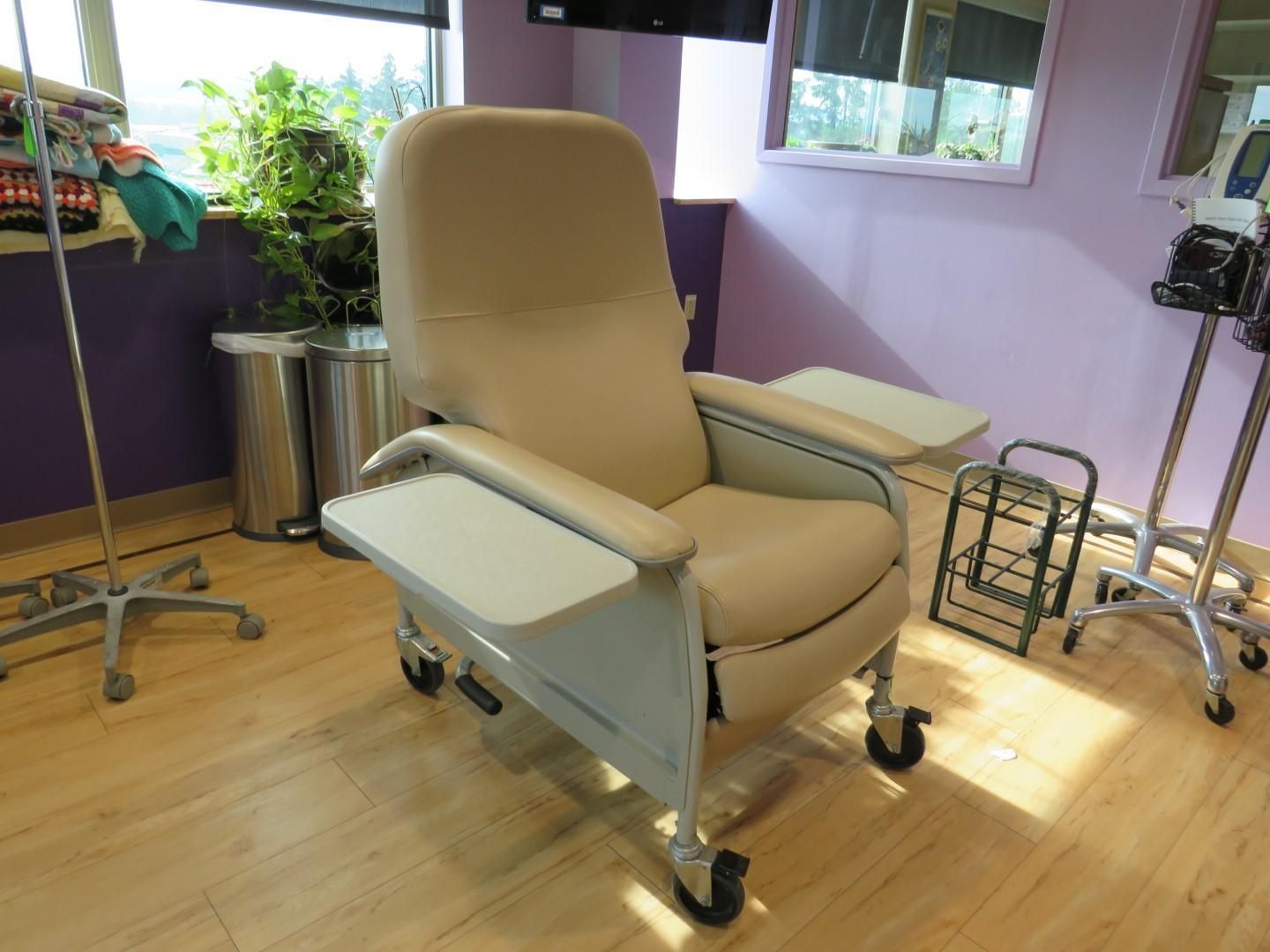 Beige medical recliner with trays in a room with purple walls, near a window and IV stands.