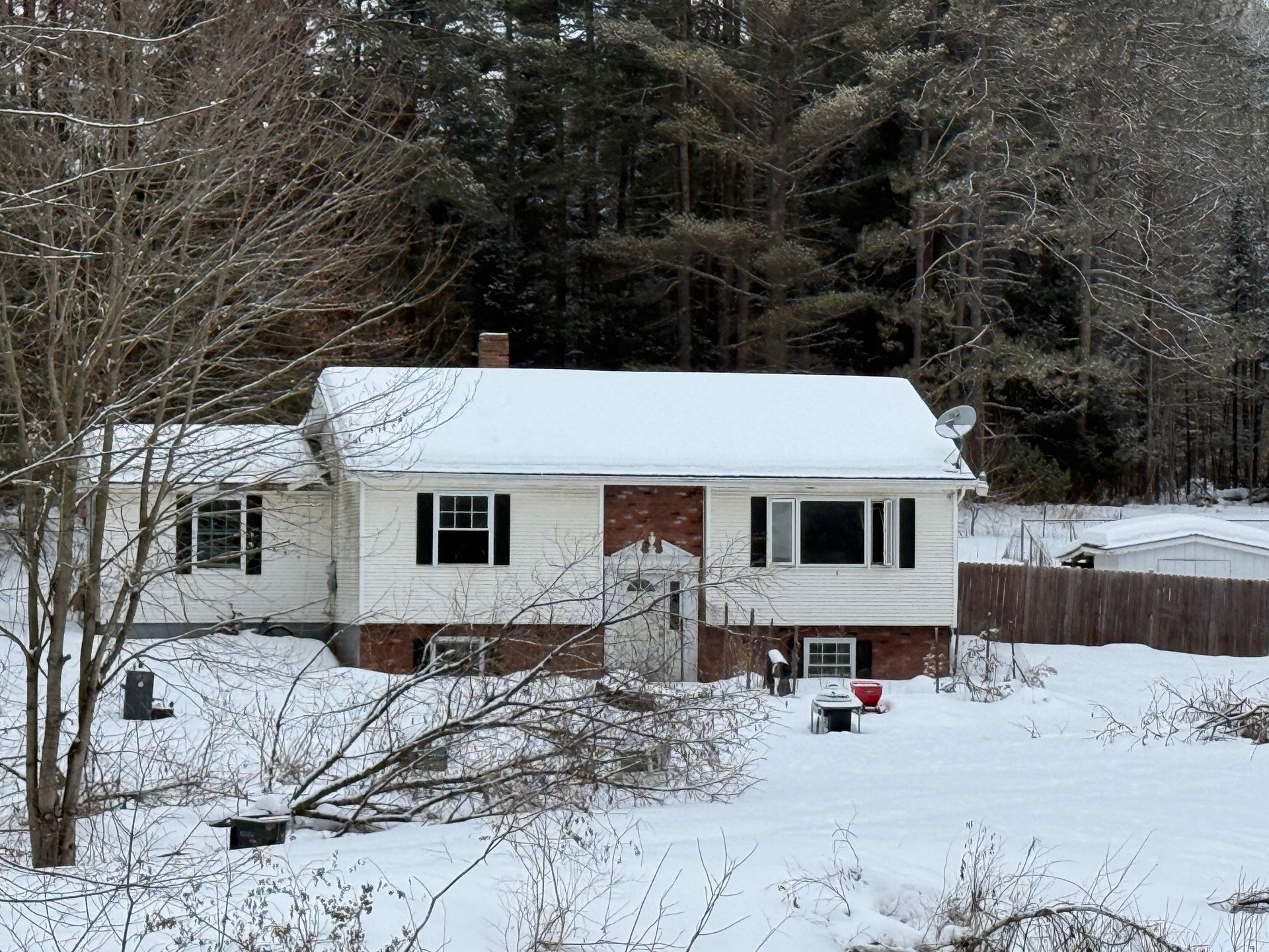 Snow-covered white house with black shutters and a brick facade, set in a snowy, wooded area.