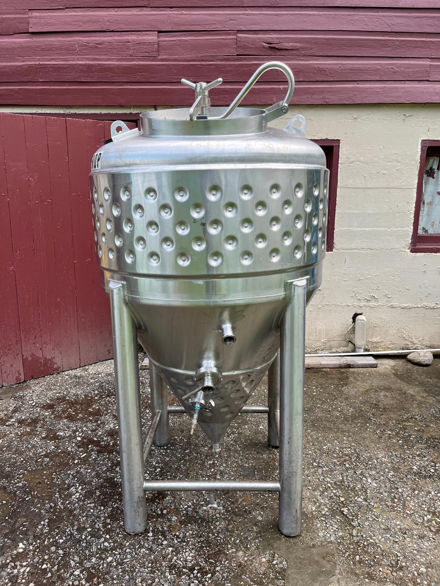 Stainless steel conical fermenter with a dimpled exterior, legs, and a red building background.