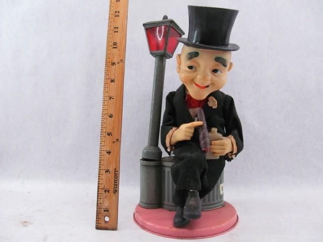 Figurine of a man in a top hat, sitting by a lamppost, pointing at a bottle.