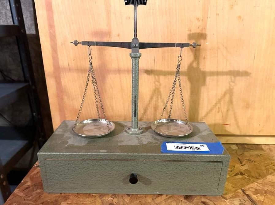 A vintage silver balance scale with two circular pans, centered on a gray box with a drawer.