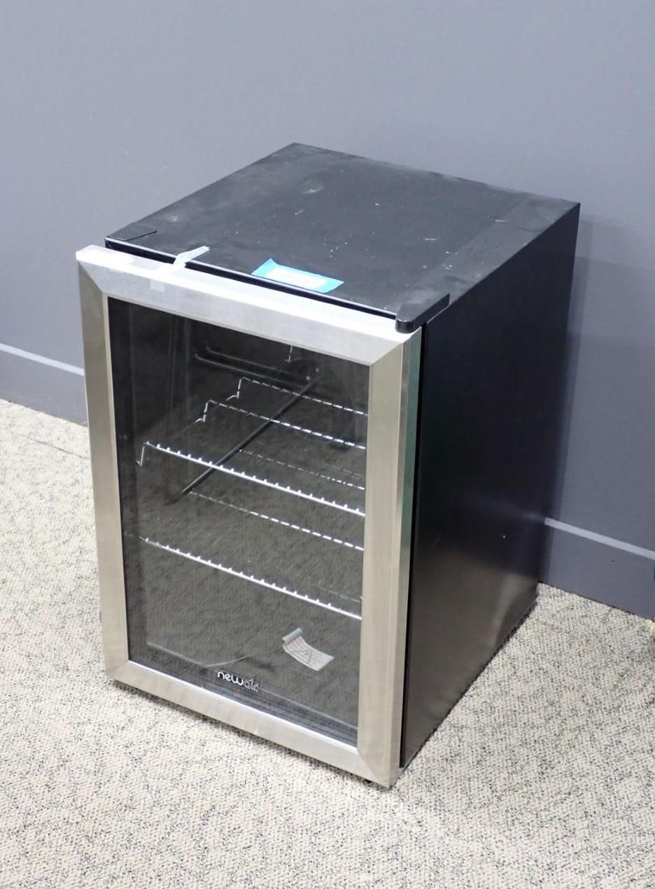 Small black and silver beverage cooler with a glass door and wire shelves, indoors.
