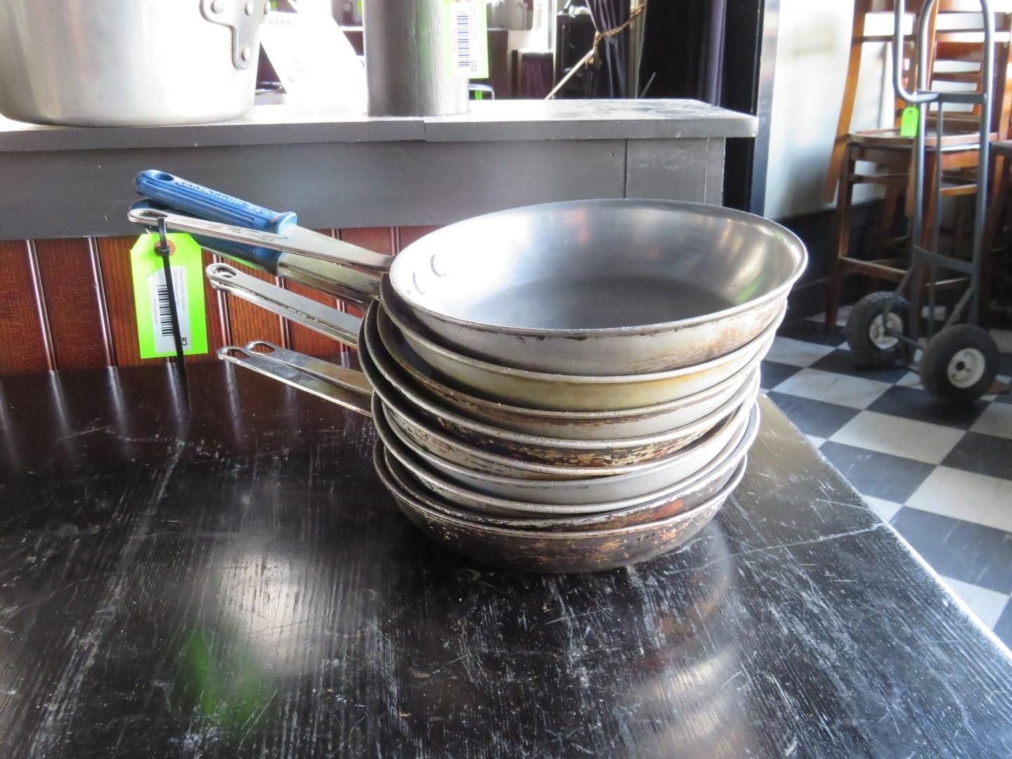 Stack of metal frying pans with blue handles on a dark table, some with price tags.