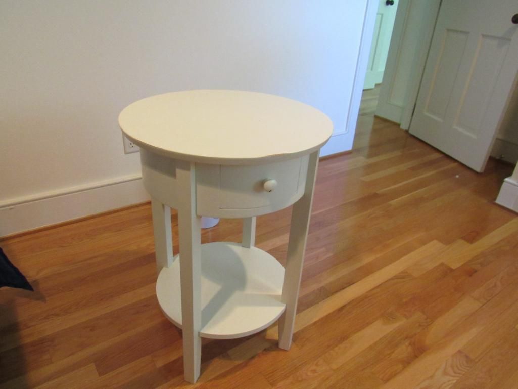 White, round side table with drawer and lower shelf on a hardwood floor.