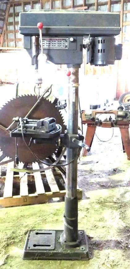 A drill press machine with a large saw blade in the background. The drill press is grey and black.