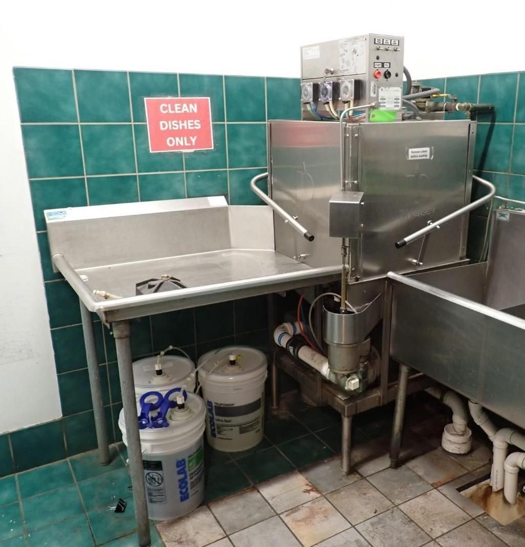 Commercial dishwasher in a kitchen setting with stainless steel tables and a sign that says 
