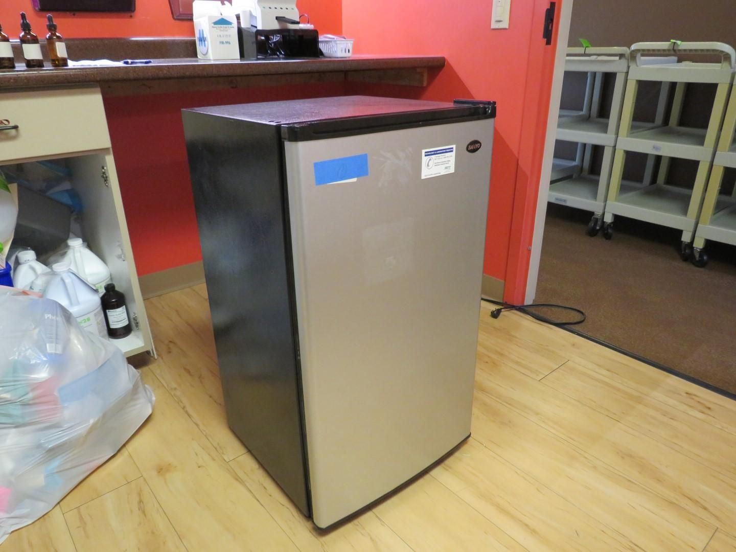 Silver mini-refrigerator against a red wall, with a wooden countertop and a doorway with shelves in the background.