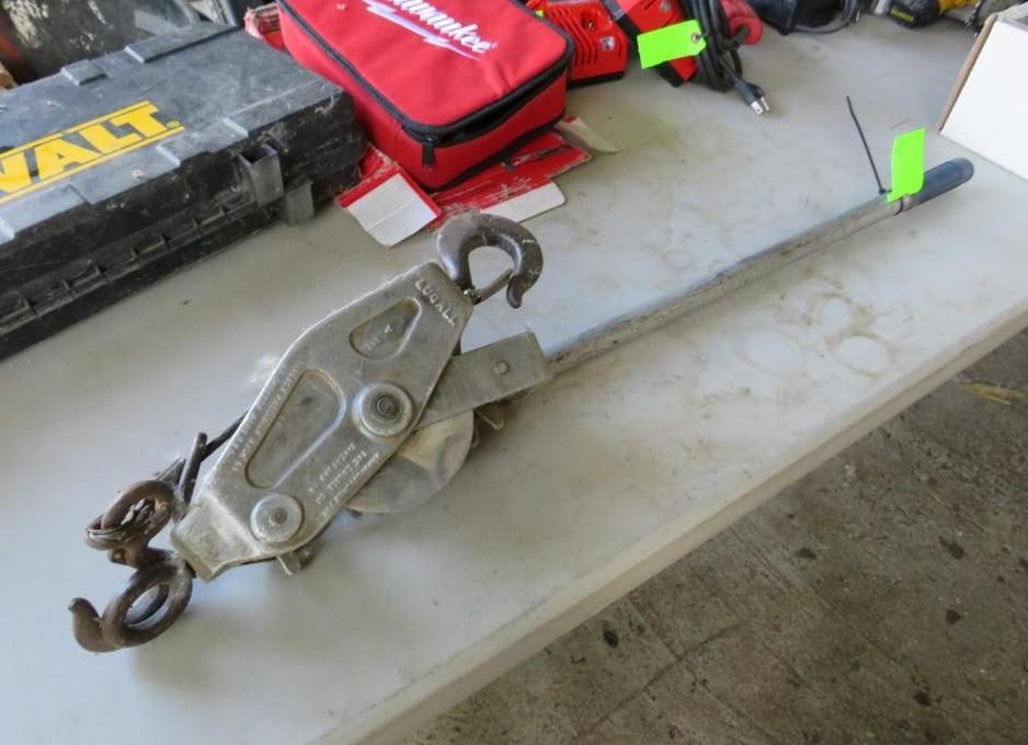 A silver come-along tool with hooks on a white table next to a black toolbox and red bag.