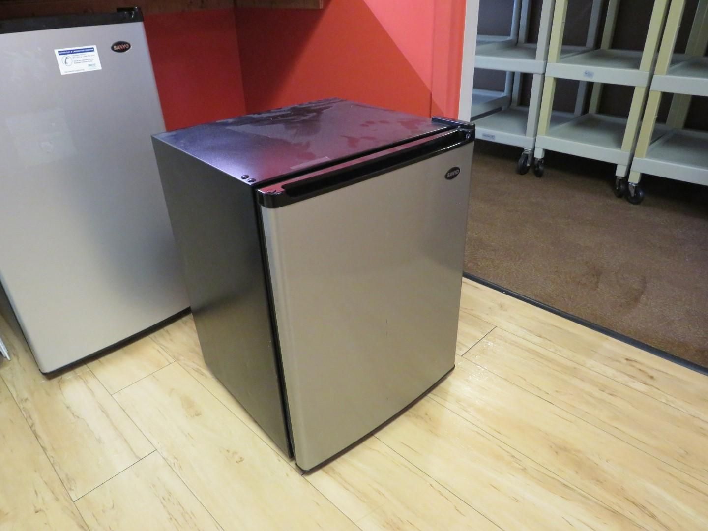 A mini-fridge in a room with a red wall and tan flooring. Another fridge is visible to the left.