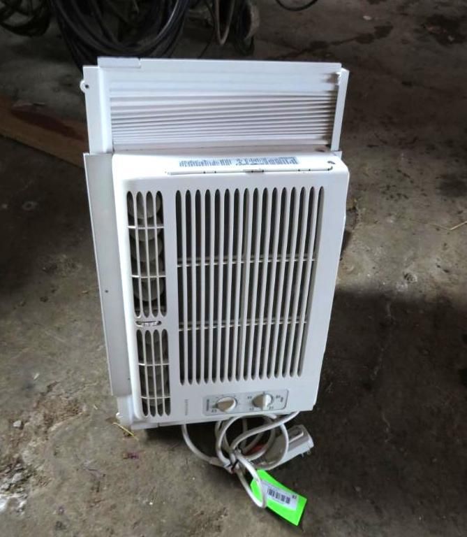 White window air conditioner with front vent and top air exhaust, sitting on a concrete floor.