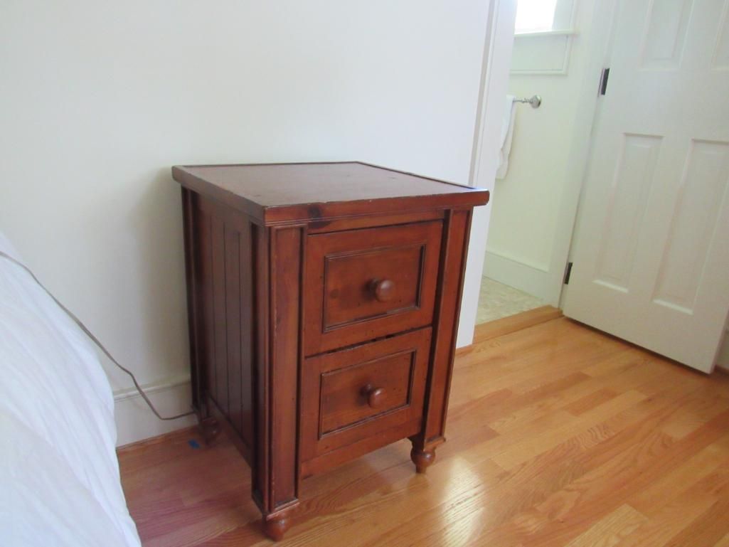 Wooden nightstand with two drawers and round knobs, brown, next to a bed and doorway.