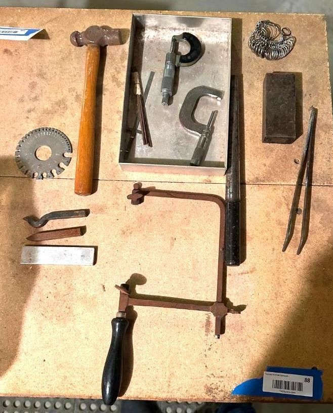 Jewelry-making tools arranged on a wooden surface: hammer, saw, files, pliers, and various metal pieces.