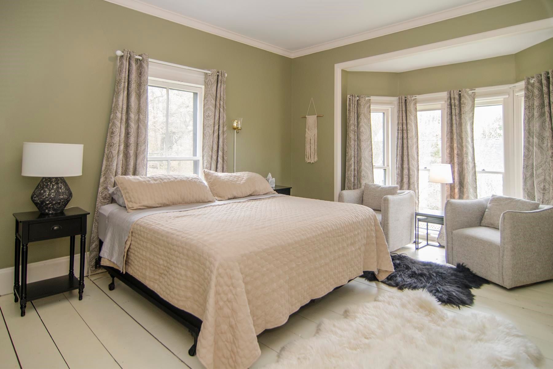 Bedroom with bed, chairs, and bay window; light green walls, pale bedding, and a fluffy rug.