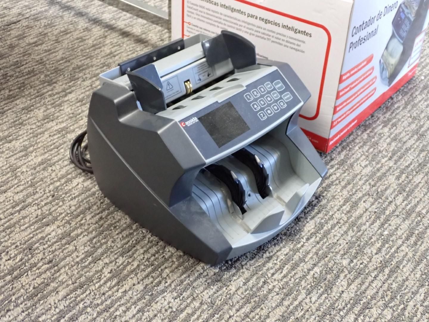 Money counting machine on a carpet next to a box. Grey and black machine with buttons and two slots.