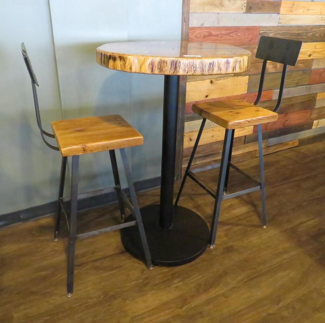 A high table with two matching stools with wooden seats and black metal frames set against a wood-paneled wall.