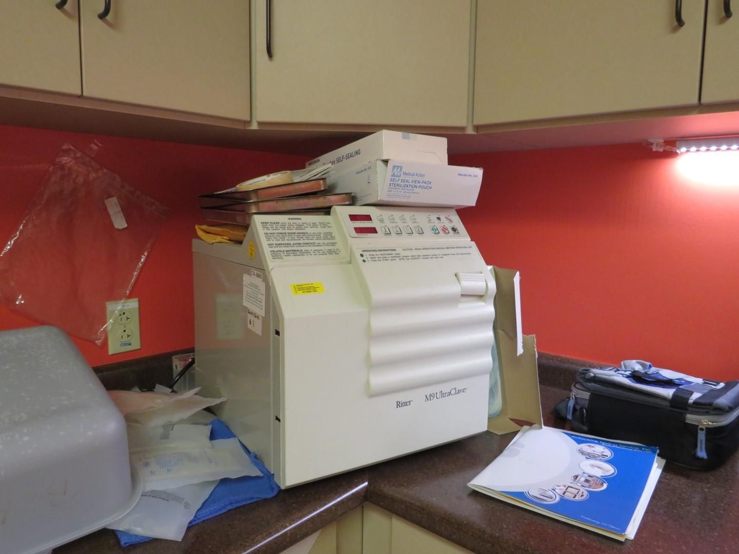 Dental equipment on a counter in a corner of a room with orange walls and white cabinets.