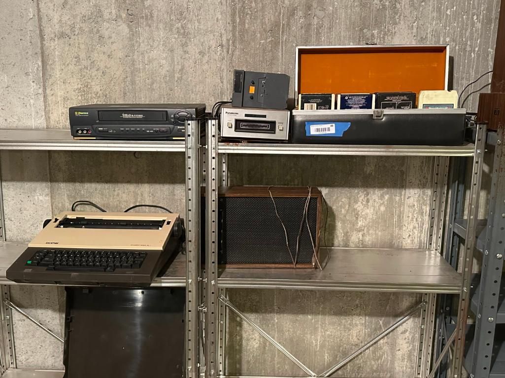 Shelves holding vintage electronics: VCR, typewriter, tape player, film equipment, and cassettes.