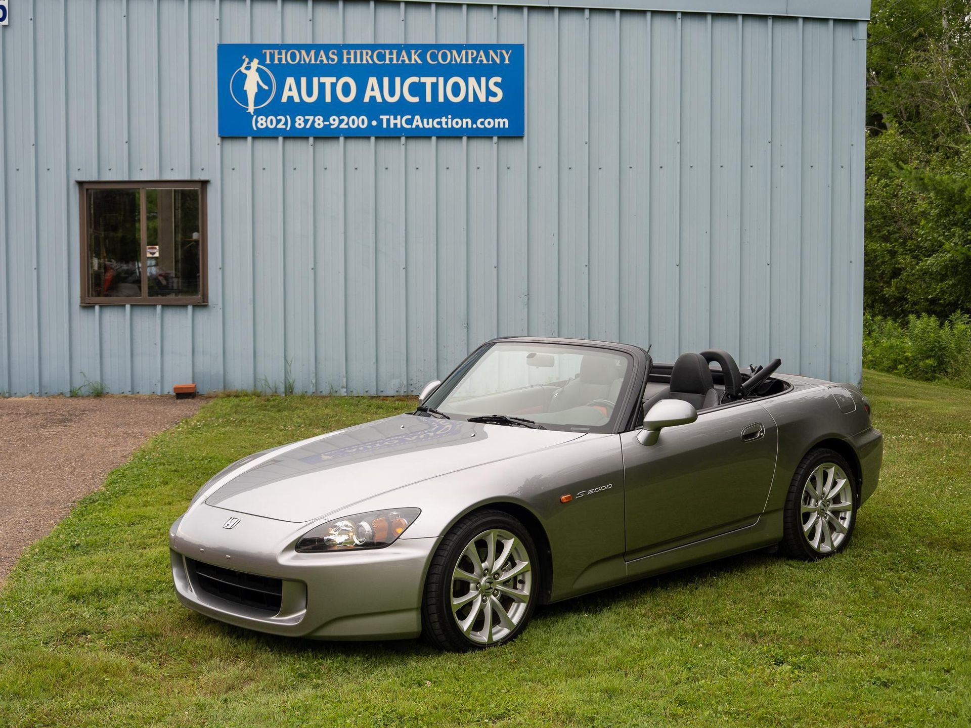 Silver Honda S2000 convertible parked on grass in front of an auto auction building.
