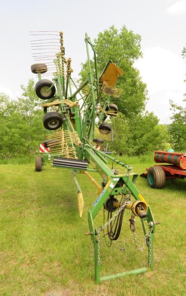 Green Krone hay rake on a grassy field, with several wheels and tines, near trees.
