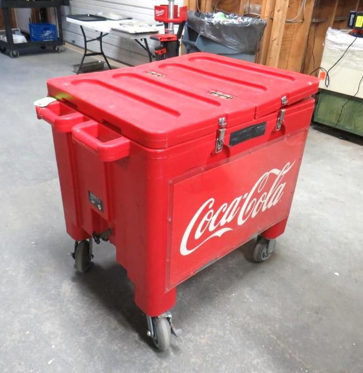 Red Coca-Cola cooler on wheels, in a workshop. It has a closed lid and metal latches.