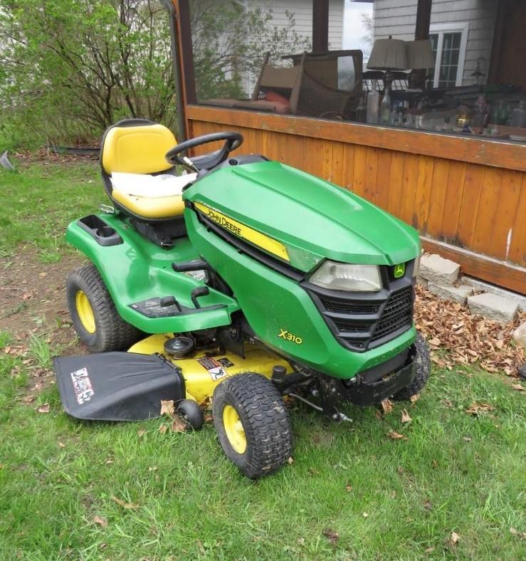 John Deere riding lawn mower on grass next to a house with a wooden porch.