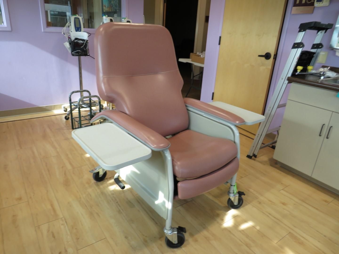 Pink medical chair with tray and armrests on wheels, in a room with medical equipment.