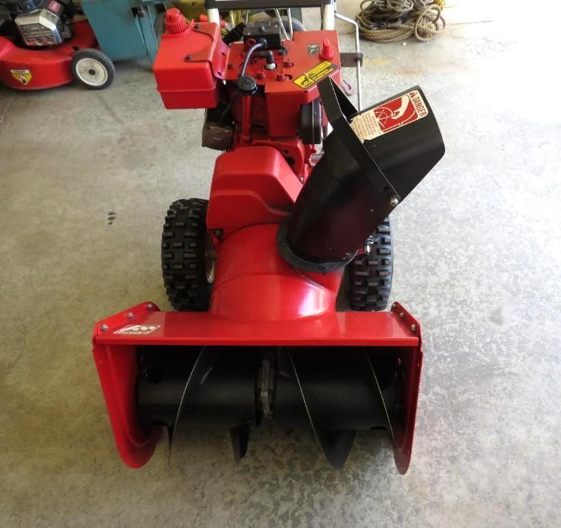 Red and black snowblower on concrete floor, with black auger blades and a black chute.