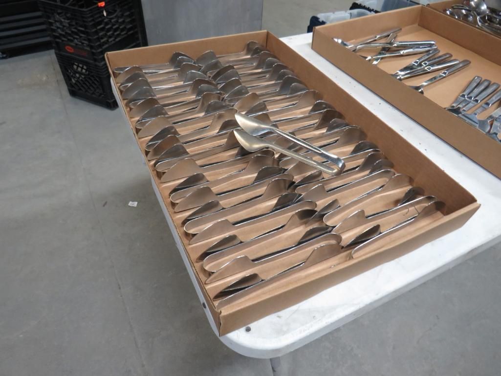 Box of silverware, including spoons and forks, and tongs, on a table.