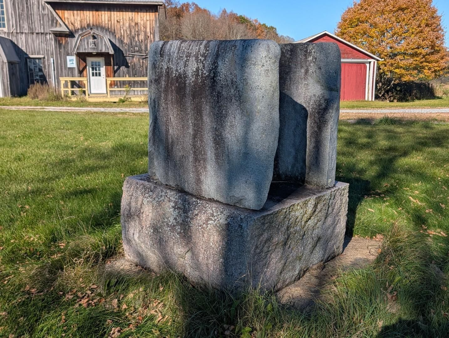 Gray stone sculpture on a grassy lawn in front of barns and autumn trees