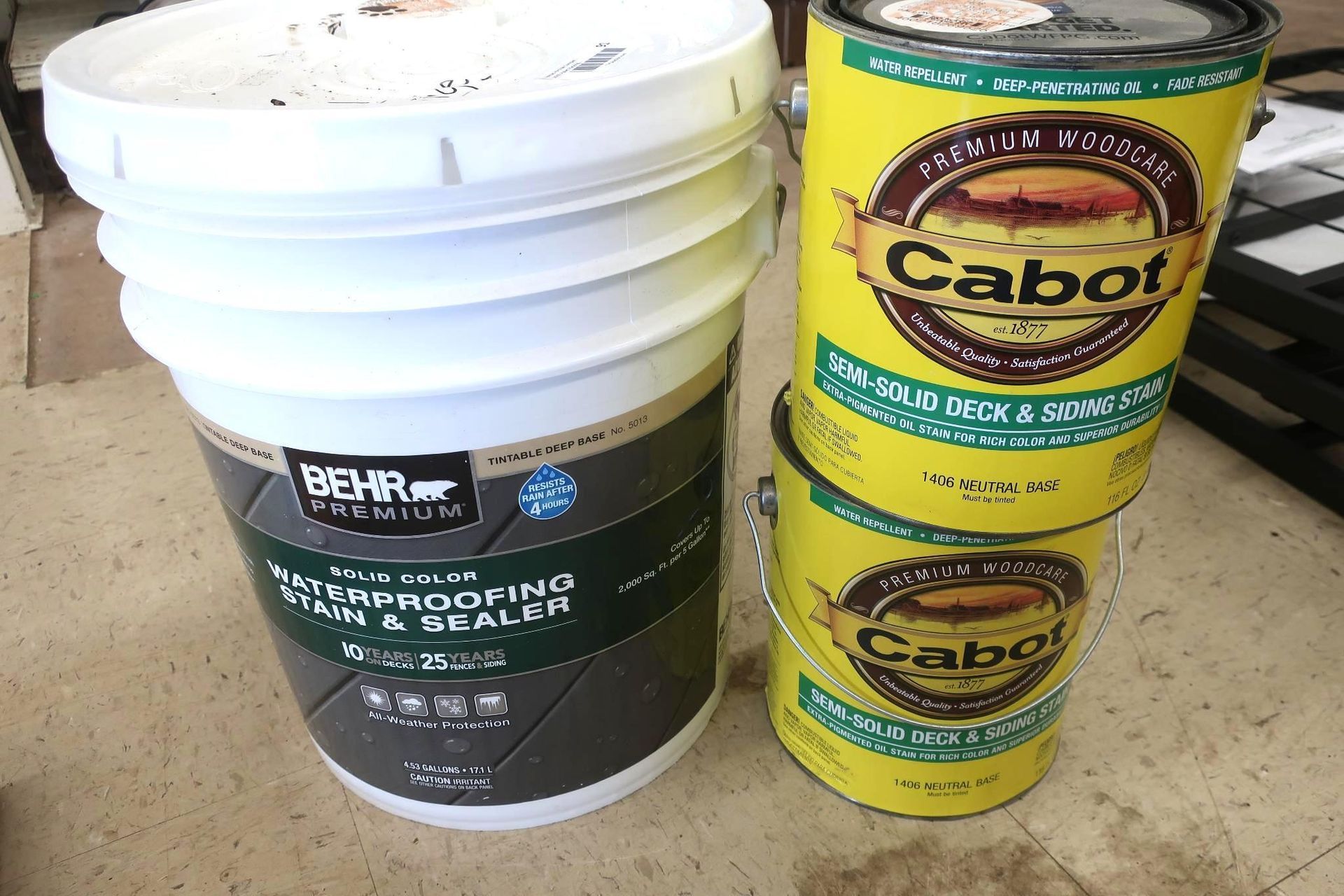Two cans of Cabot stain and one can of Behr waterproofing stain on a surface.