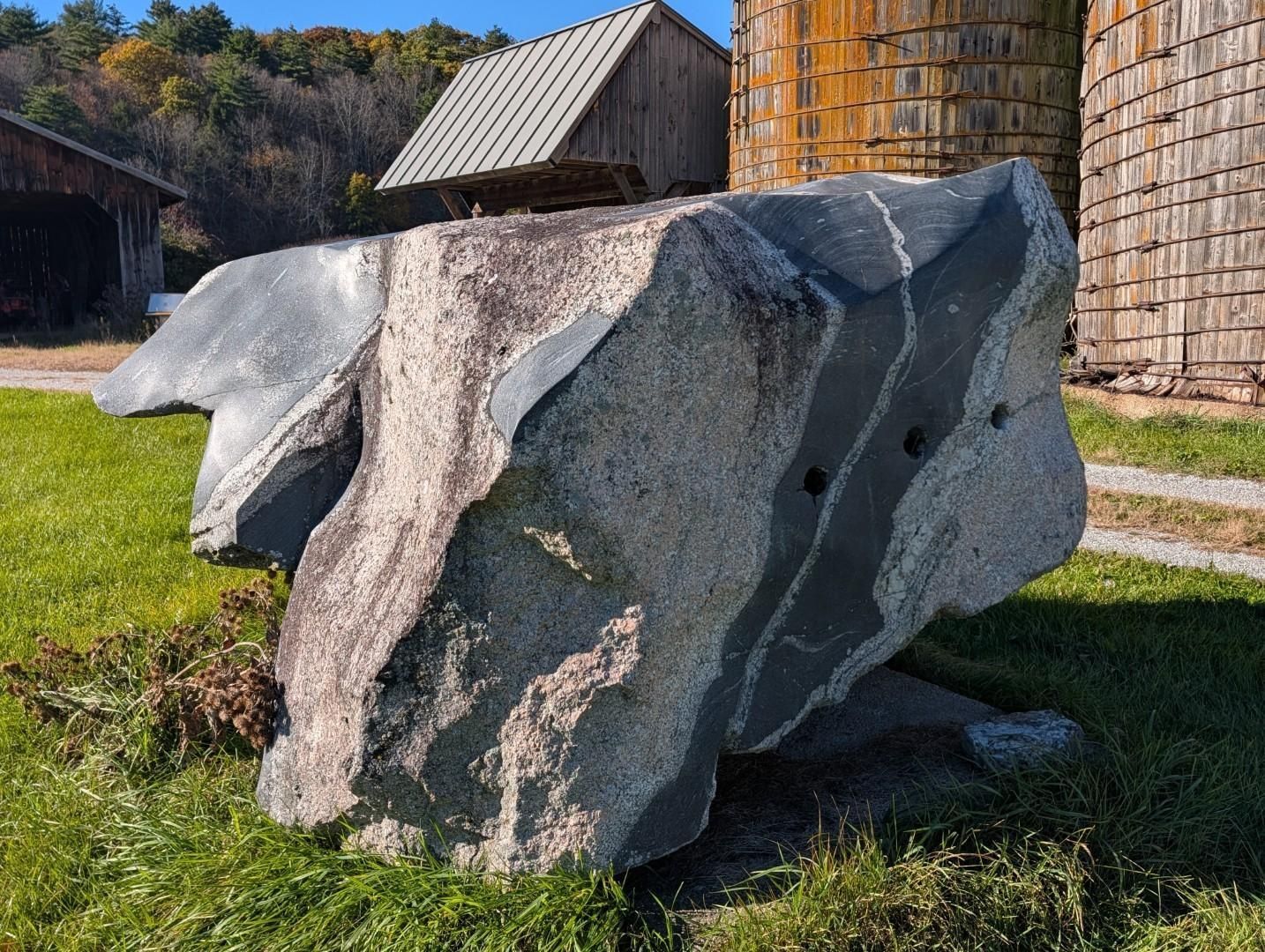 Large gray boulder on grass beside a farm building and wooden silo under a sunny sky