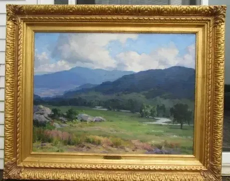 Landscape painting of a valley with mountains in the background, set in a gold ornate frame.