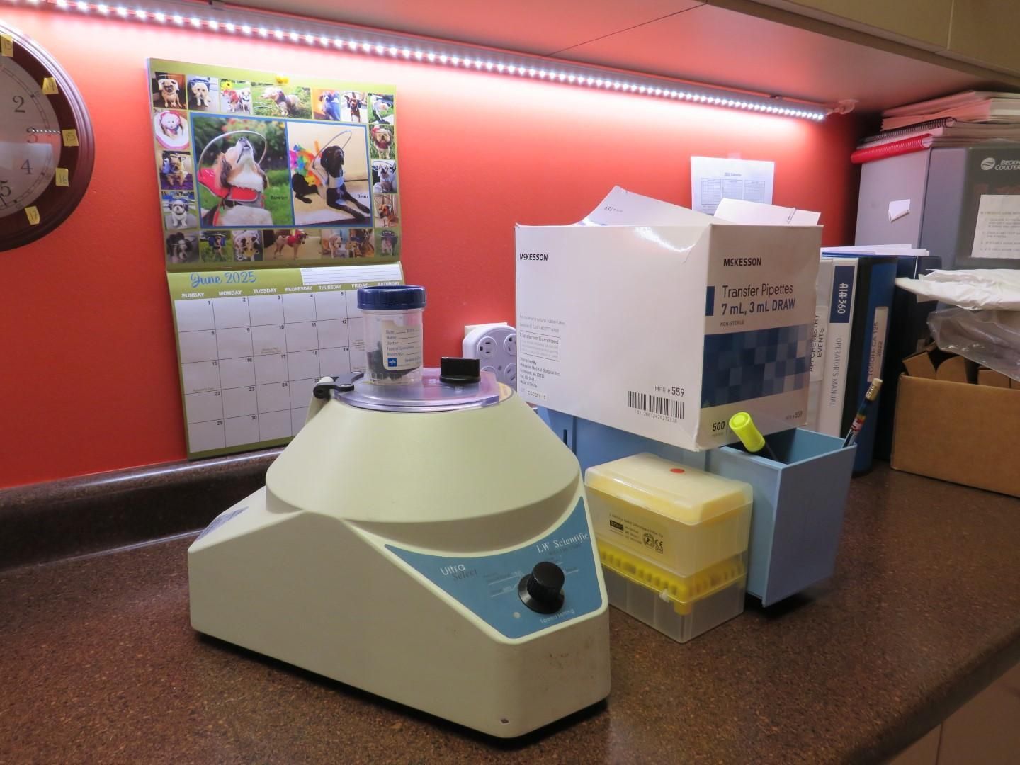A laboratory vortex mixer with vials, boxes, and other equipment on a counter under a red wall.